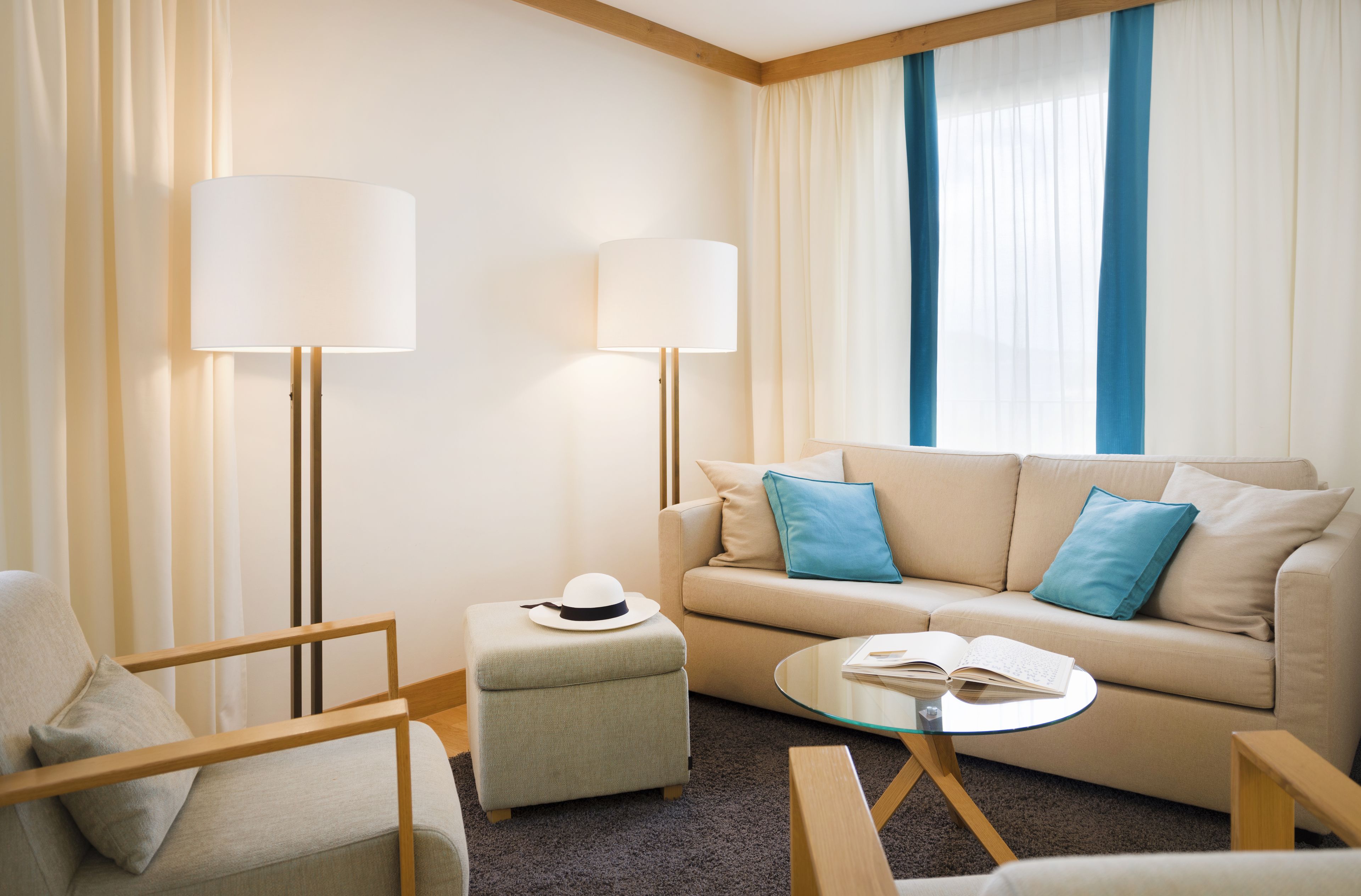 Una habitación de hotel moderna con sofá, sillón, reposapiés y una mesa de centro de vidrio.