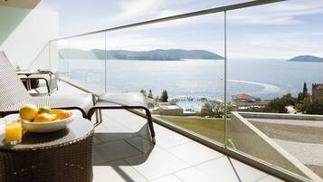 A balcony with wicker furniture, a glass table with fruit, and a view of the sea and distant hills.