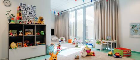 A brightly lit room with a large window, a bookshelf filled with toys, and a rug with various plush toys.