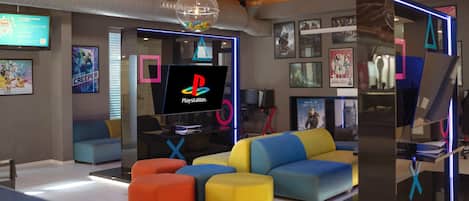 Una moderna area giochi con sedute colorate, una TV PlayStation e diversi poster.
