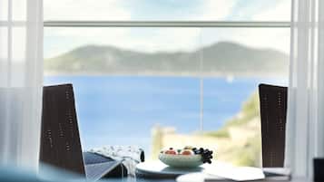 A dining table with a bowl of fruit and a napkin, positioned by a window with a view of the sea and hills.