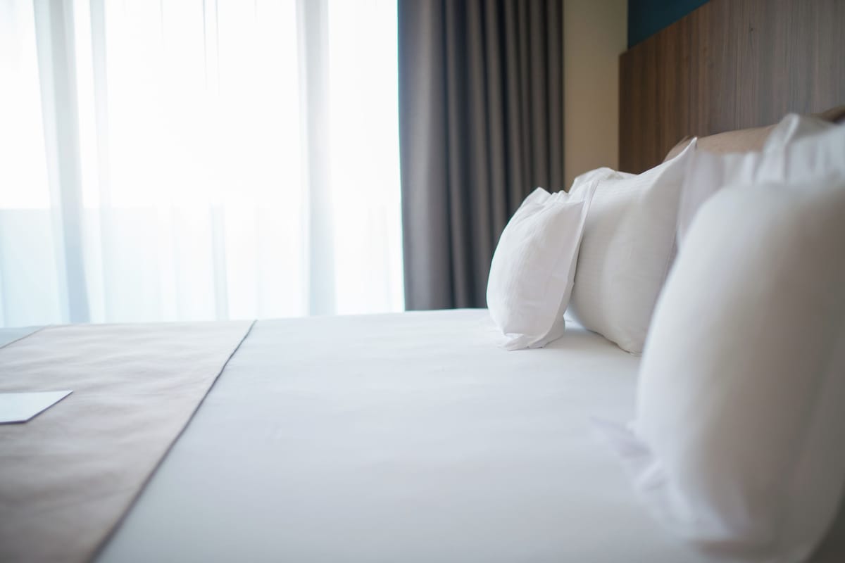 A neatly made bed with white linens and pillows, a wooden headboard, and a window with curtains.