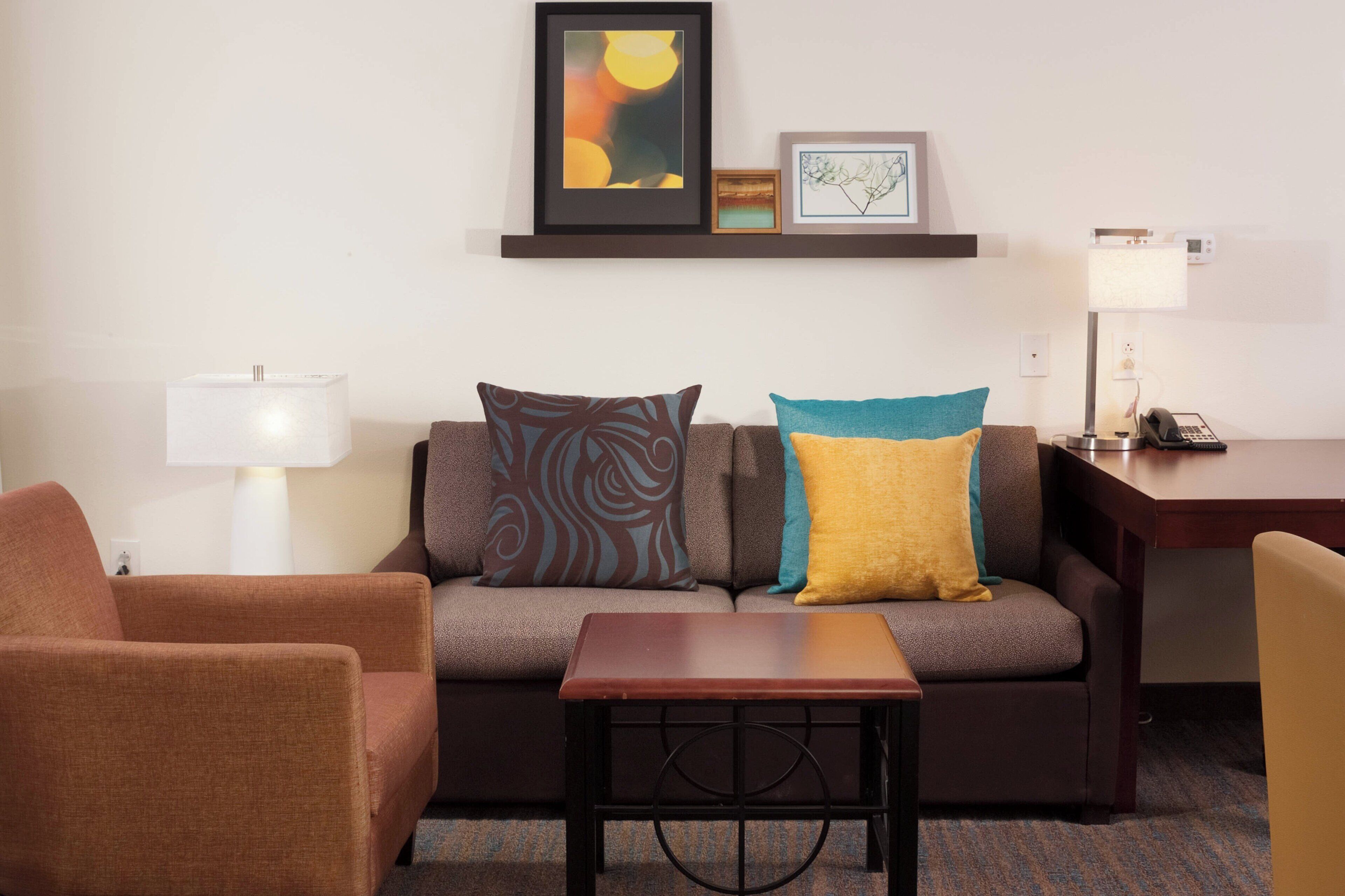 Une chambre d’hôtel moderne, dotée d’un canapé, d’un fauteuil et d’une petite table. On y trouve un bureau avec une lampe et un téléphone. La chambre présente un aménagement simple et épuré, avec un décor minimaliste.