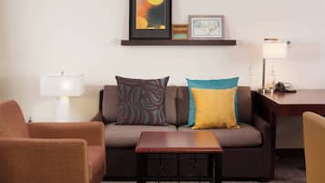 Une chambre d’hôtel moderne, dotée d’un canapé, d’un fauteuil et d’une petite table. On y trouve un bureau avec une lampe et un téléphone. La chambre présente un aménagement simple et épuré, avec un décor minimaliste.