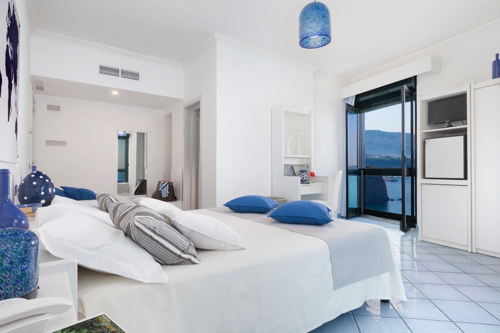 Une chambre à coucher moderne avec un lit, des oreillers, une lampe suspendue bleue, une armoire et une vue sur la mer.