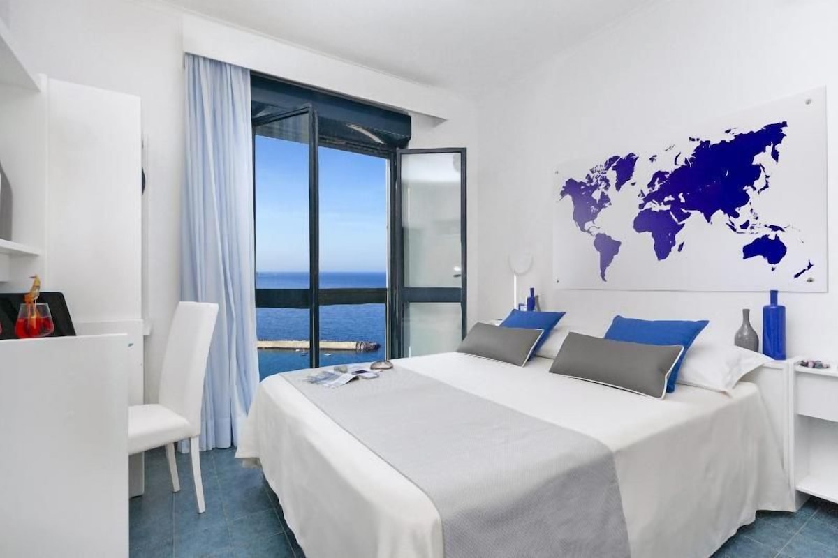 A modern hotel room with a large bed, a world map on the wall, a view of the sea, and a desk with a chair.