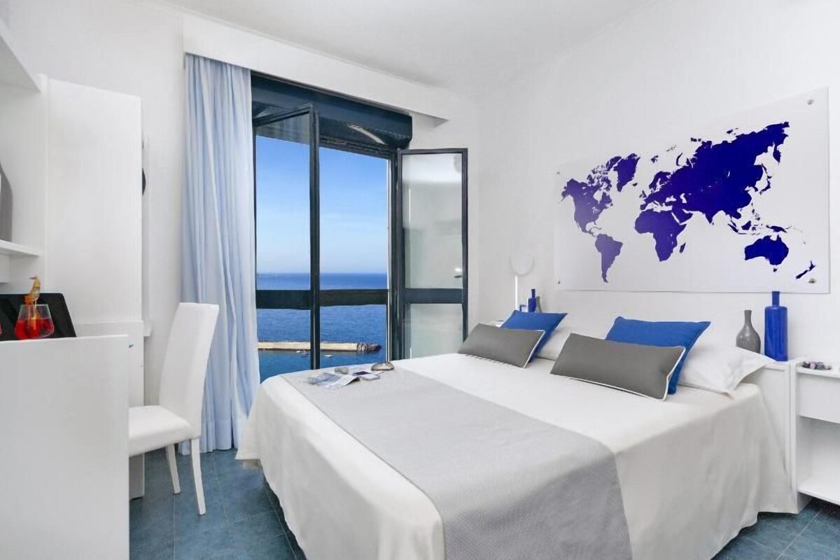 A modern hotel room with a large bed, a world map on the wall, a view of the sea, and a desk with a chair.