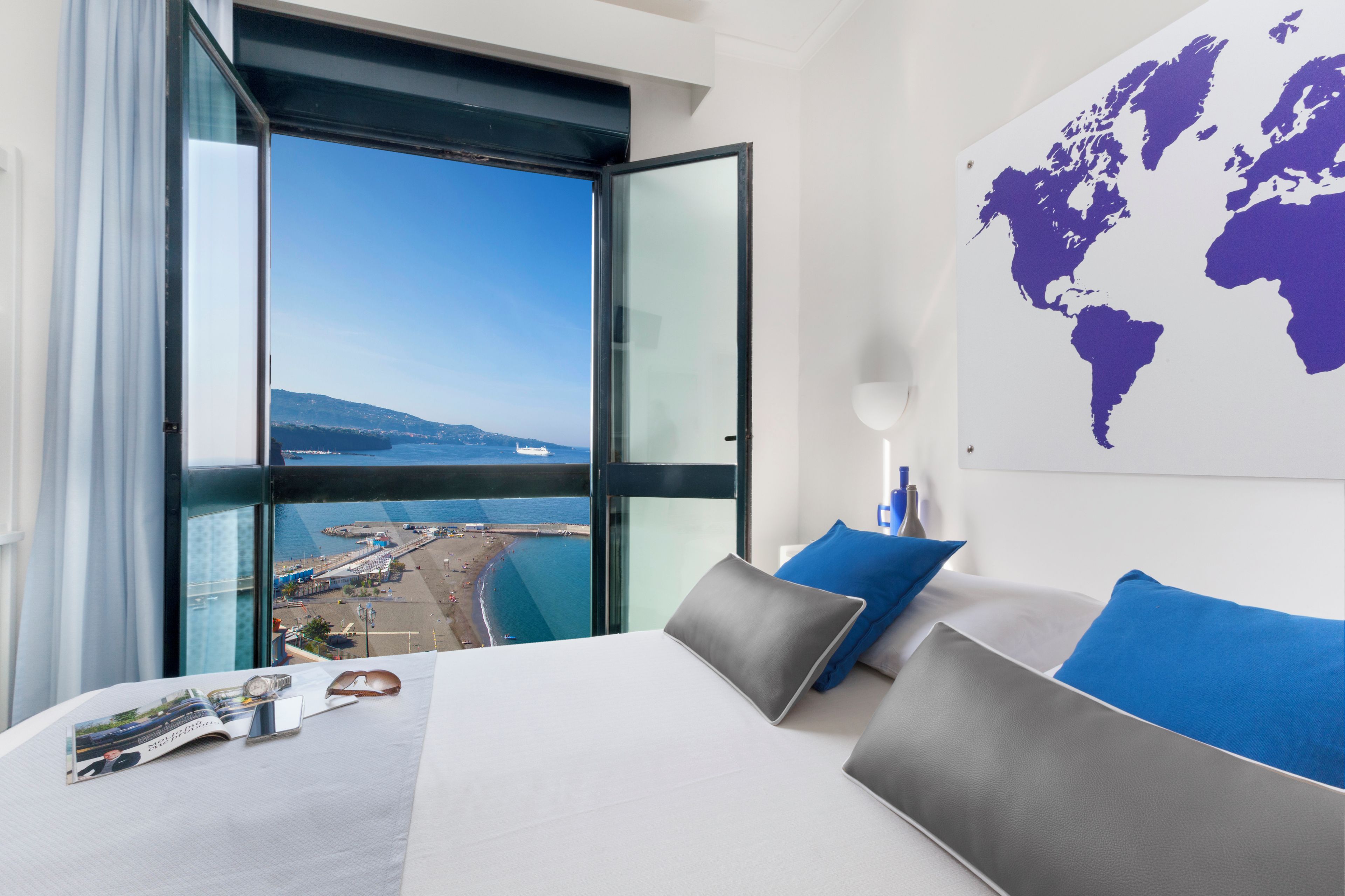 A hotel room with a large bed, a world map on the wall, and a view of a coastal area.