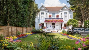 A two-story white building with a balcony, surrounded by a well-maintained garden with colorful flowers and a wooden fence.