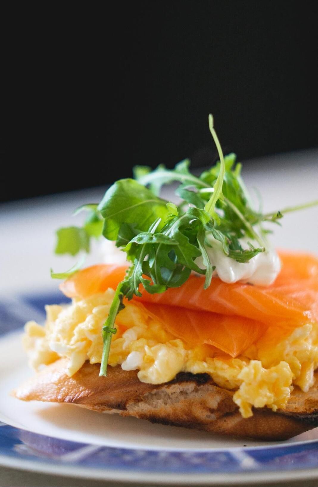 A plate with a slice of bread topped with scrambled eggs, smoked salmon, and a garnish of arugula and microgreens.