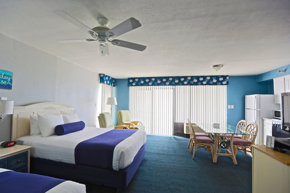 Habitación de hotel con cama, escritorio y silla. Cuenta con ventilador de techo, ventana con persianas y televisor.