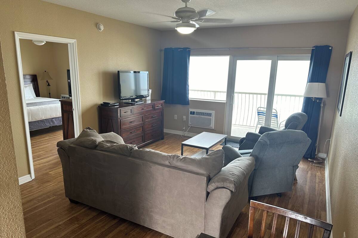 Una sala de estar con un sofá, sillones, una mesa de centro, un televisor en un aparador y un balcón con cortinas.