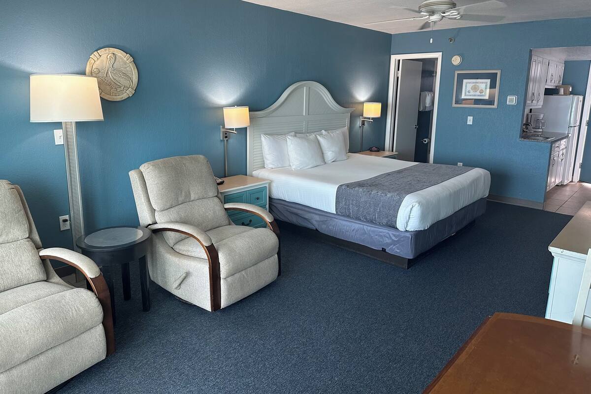 Habitación de hotel con cama, dos sillas, mesita de noche, lámpara, ventilador de techo y kitchenette.