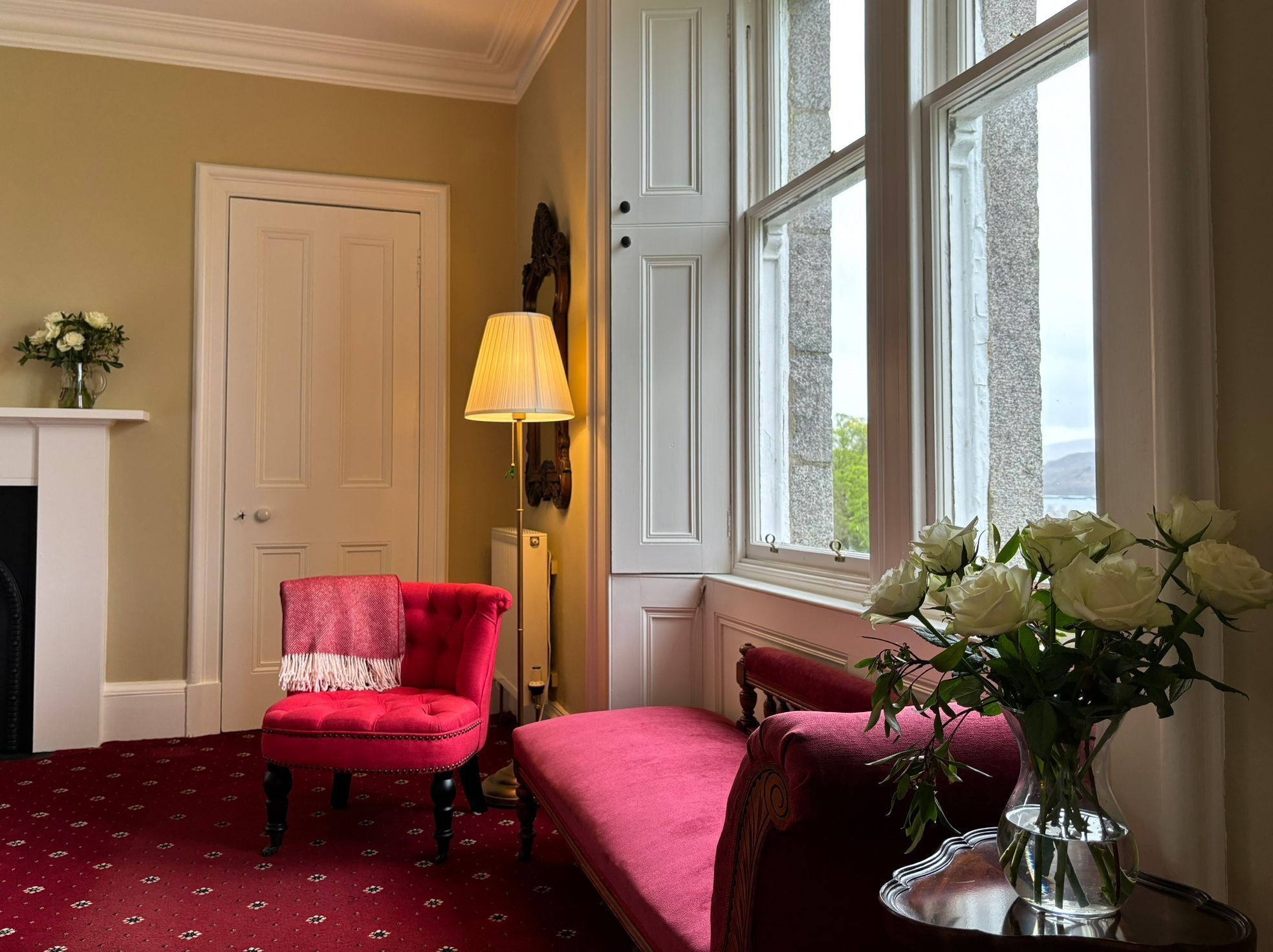A room with a red armchair, a matching bench, a lamp, and a vase of white flowers.