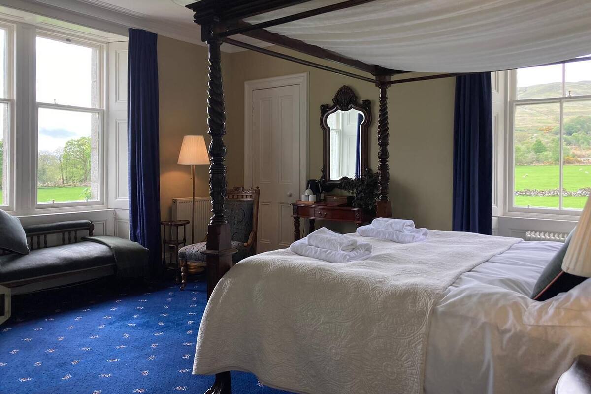 A four-poster bed with white linens, a bedside table with a lamp, and a view of a green landscape through large windows.
