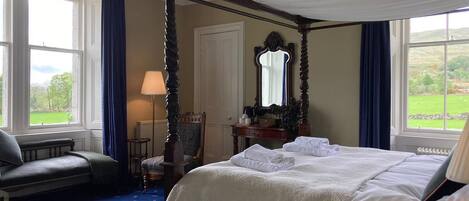 A four-poster bed with white linens, a bedside table with a lamp, and a view of a green landscape through large windows.