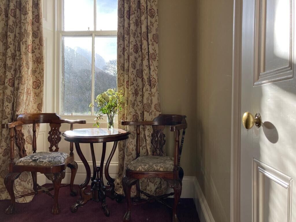 Ein Zimmer mit einem großen Fenster, zwei Holzstühlen, einem kleinen runden Tisch und einer Tür mit Messingknauf.