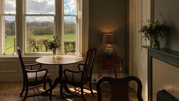 Un comedor con mesa y sillas, una lámpara y vistas a un paisaje verde a través de amplios ventanales.