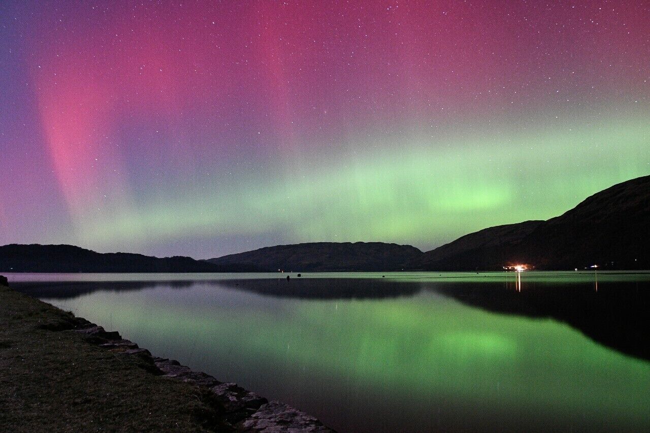 A nighttime view of a calm lake with a mountain in the background and the Northern Lights in the sky.