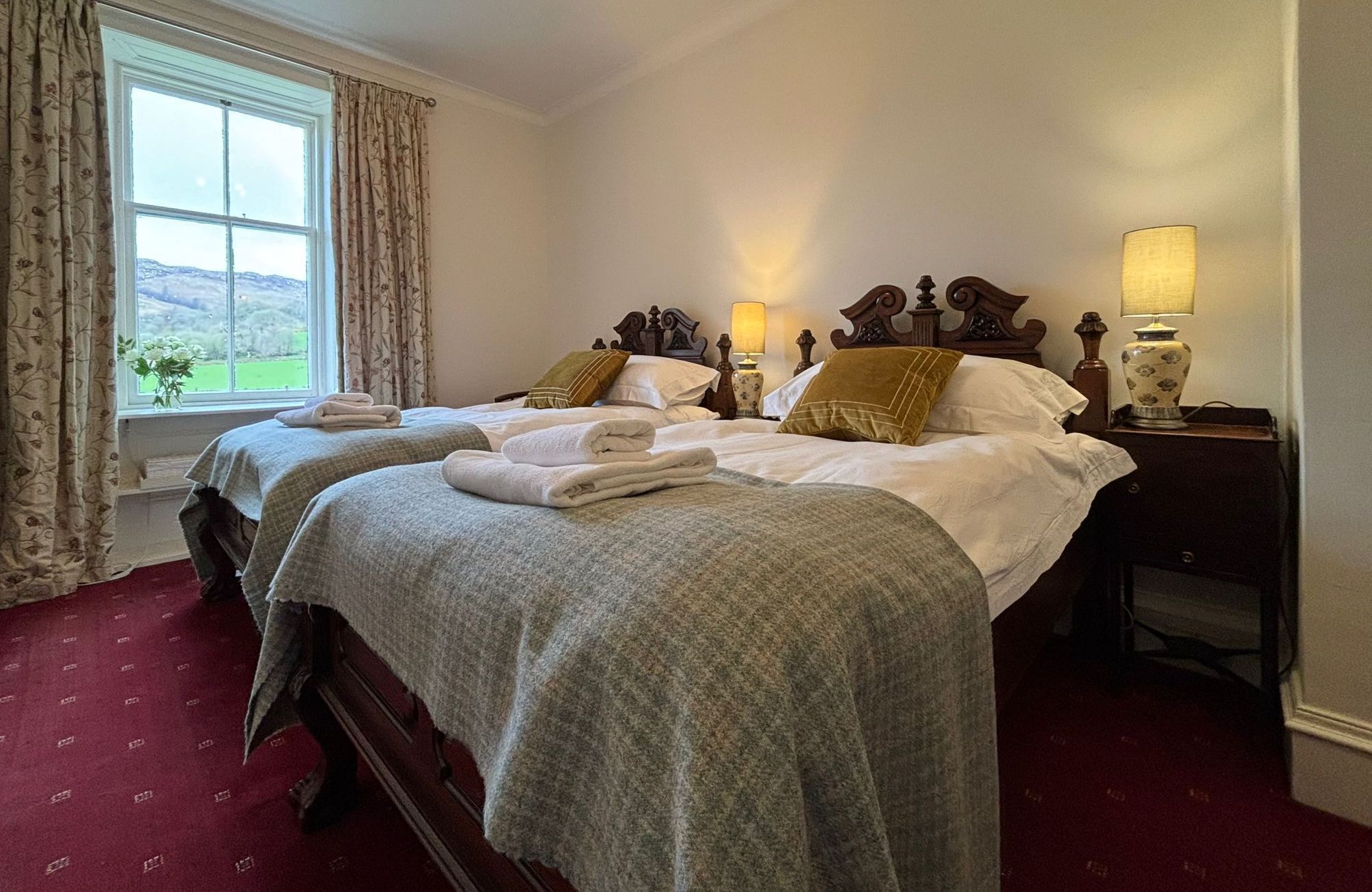 A hotel room with two beds, a window with curtains, a lamp on the nightstand, and a view of the outdoors.
