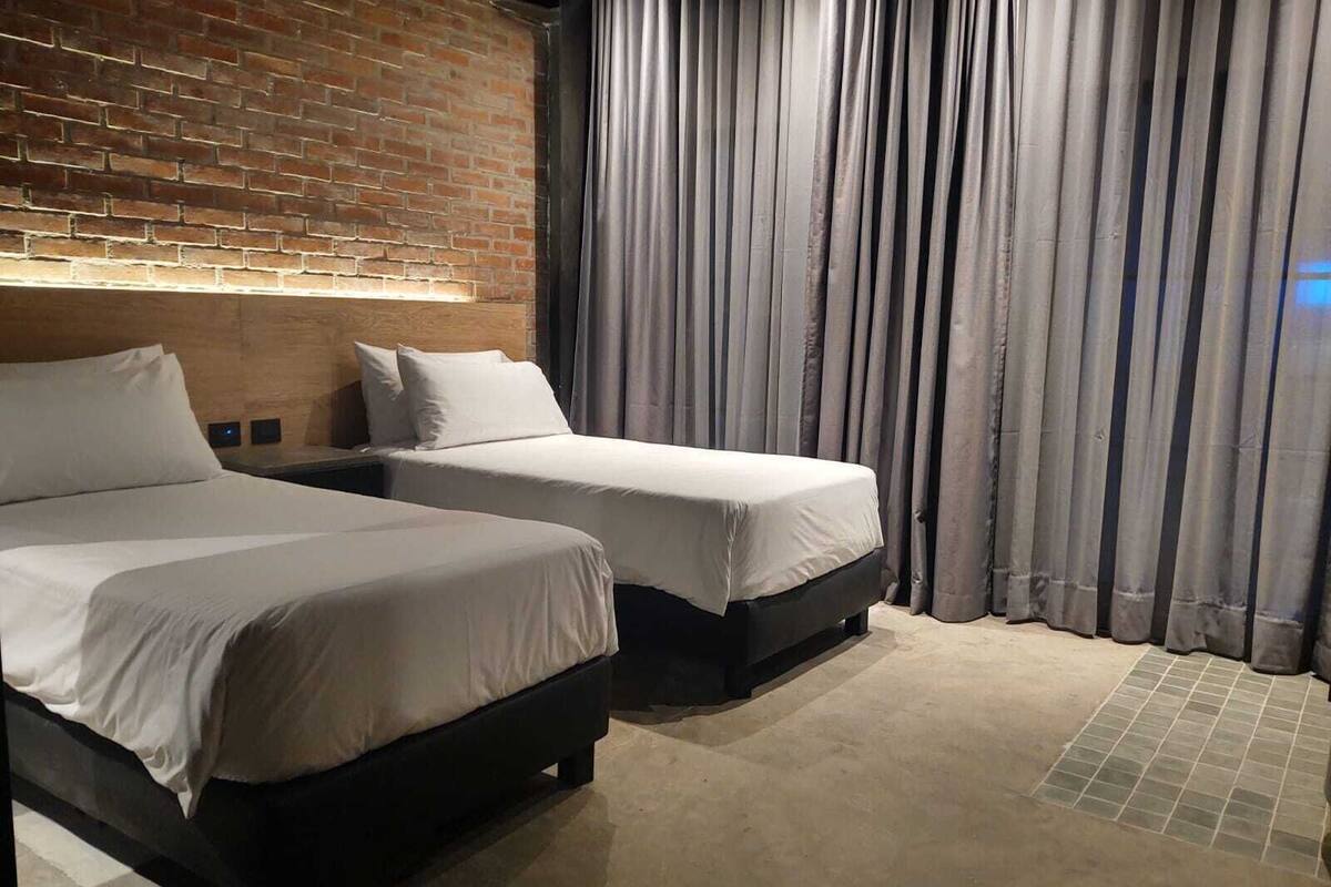 Habitación de hotel con dos camas, una pared de ladrillo y una cortina.