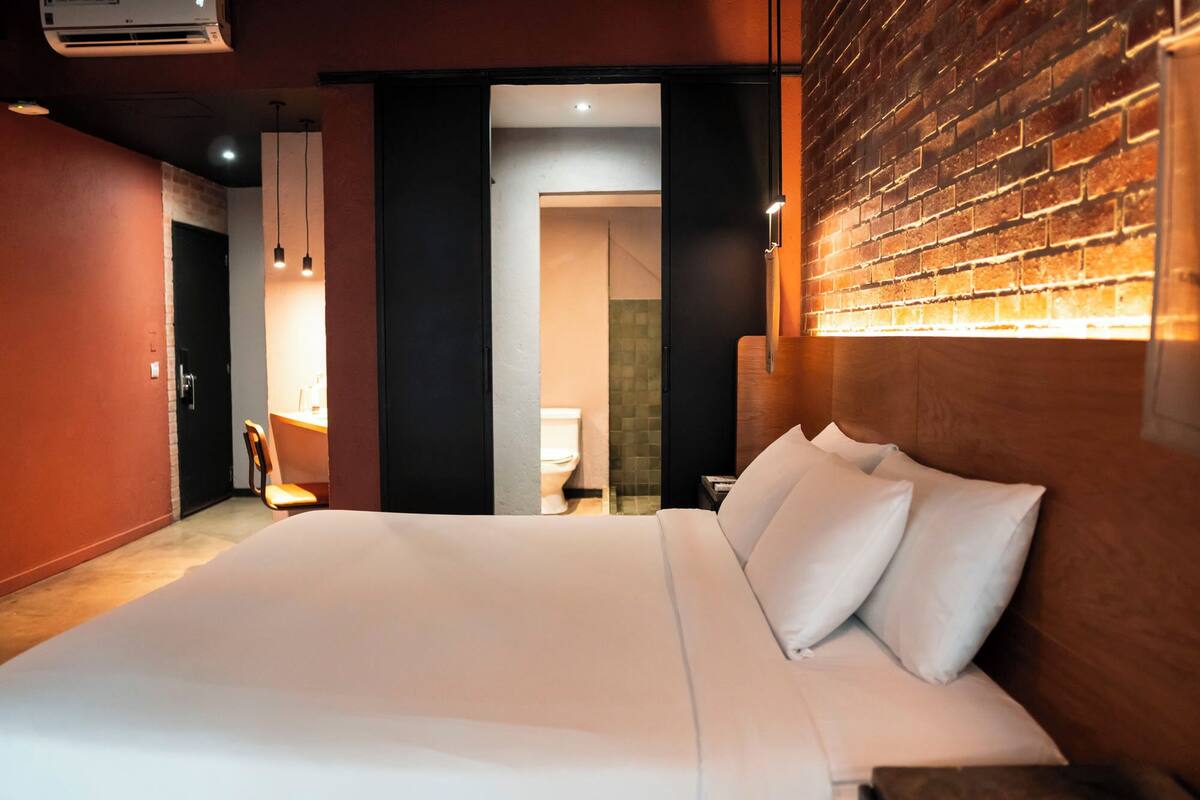 Habitación de hotel con una cama grande, cabecera de madera, pared de ladrillo y baño visible a través de una puerta abierta.