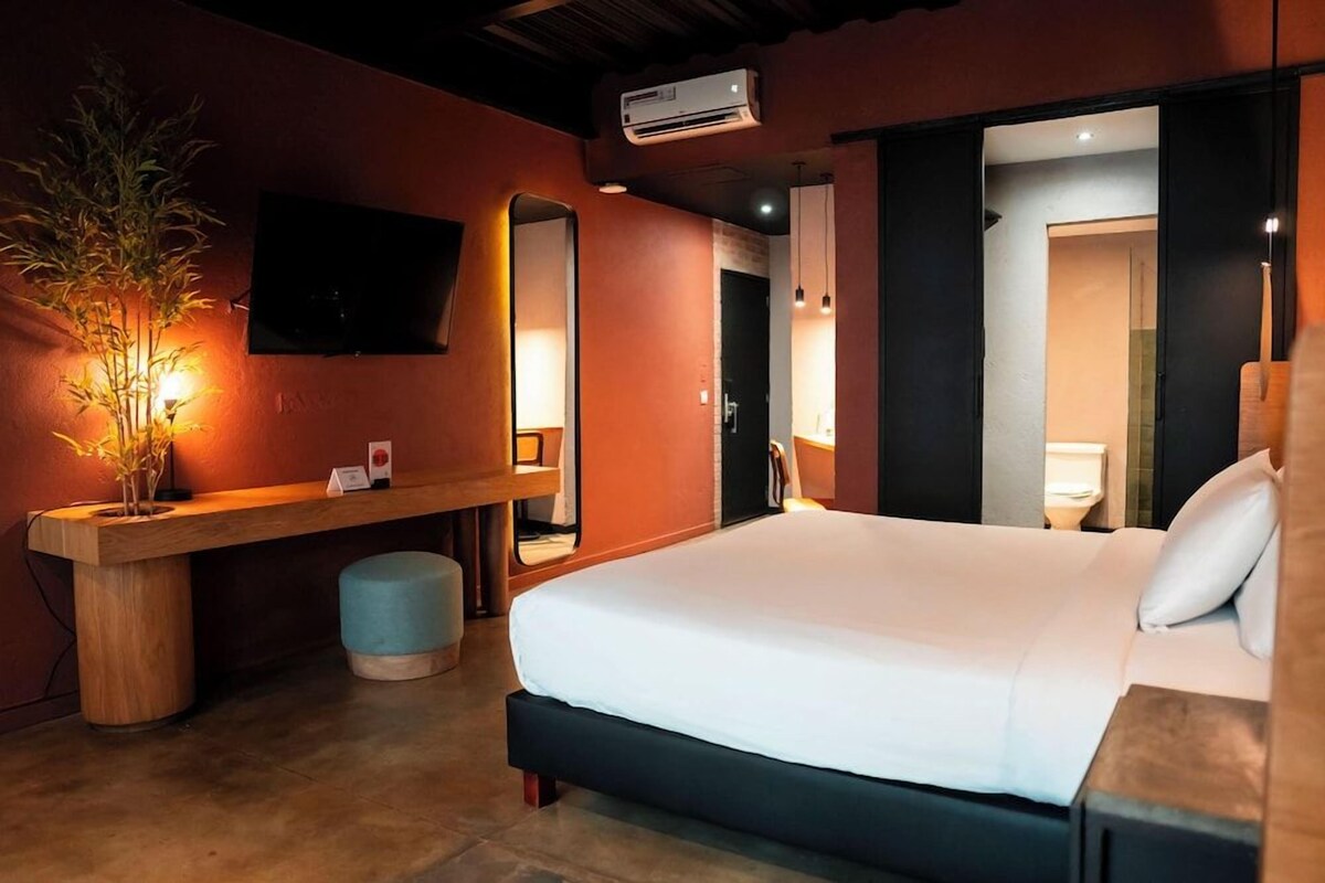 A hotel room with a bed, a desk, a mirror, a TV, and an air conditioning unit.
