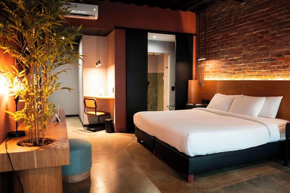 A modern hotel room with a large bed, a desk with a plant, a chair, and a brick wall.