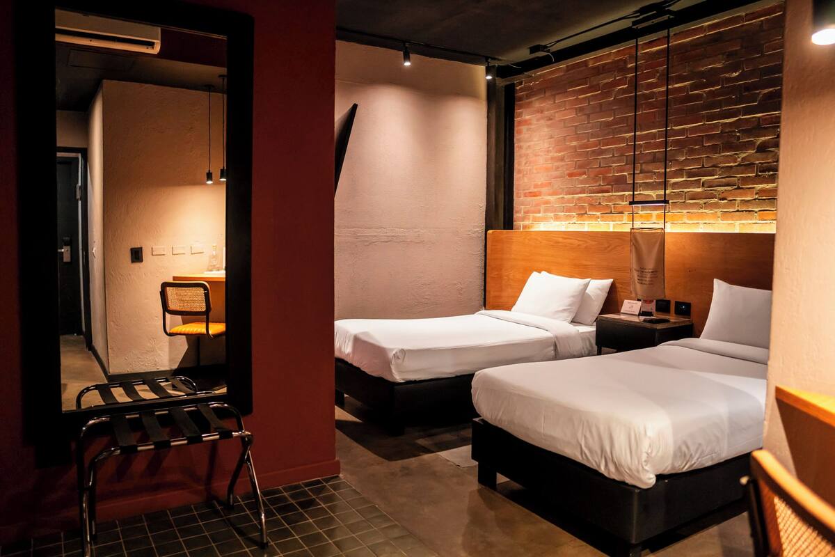 Habitación de hotel con dos camas, una silla, una mesita de noche y una pared de ladrillo.