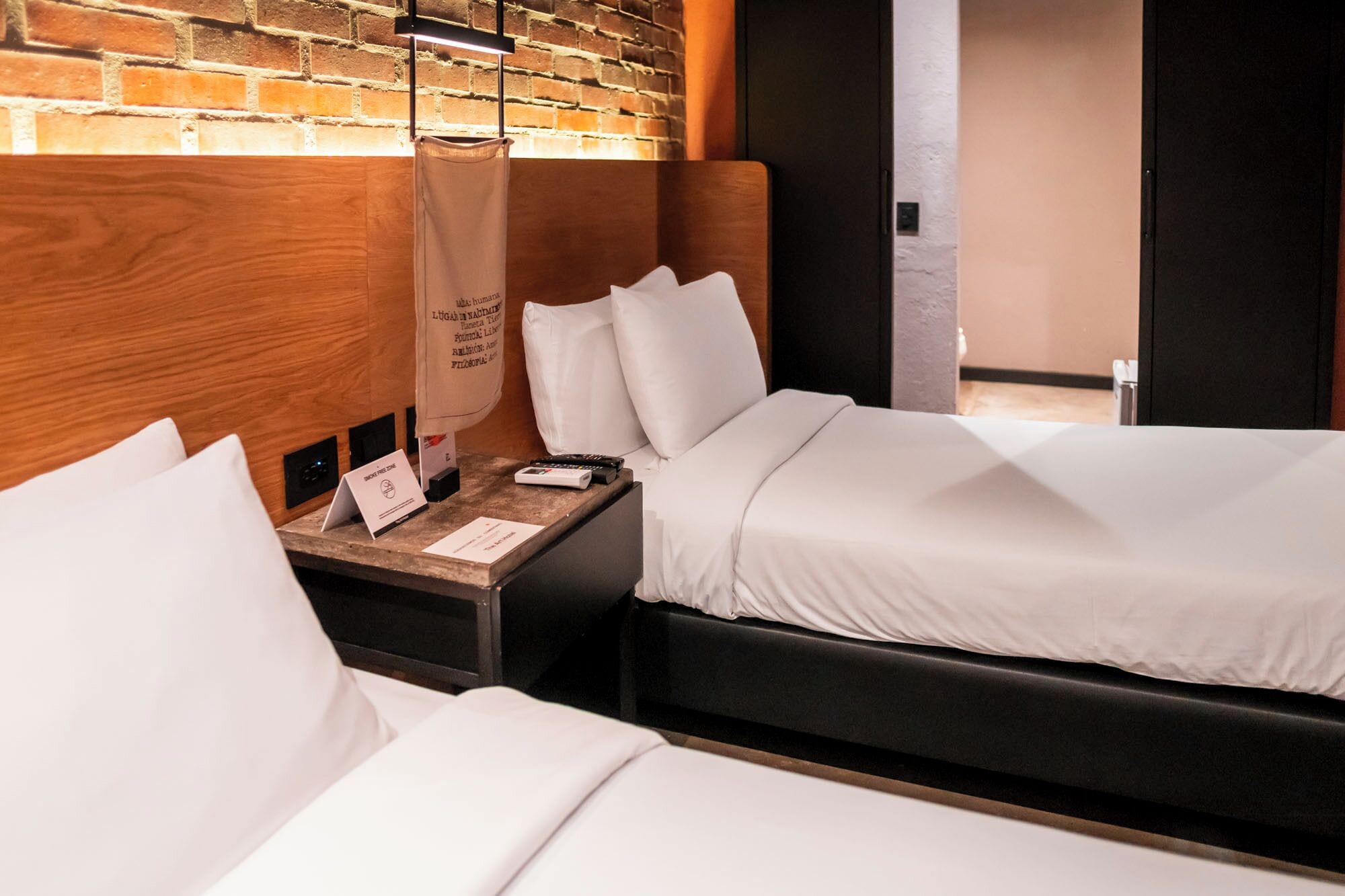 A hotel room with two beds, a wooden headboard, a nightstand with a lamp, and a brick wall.