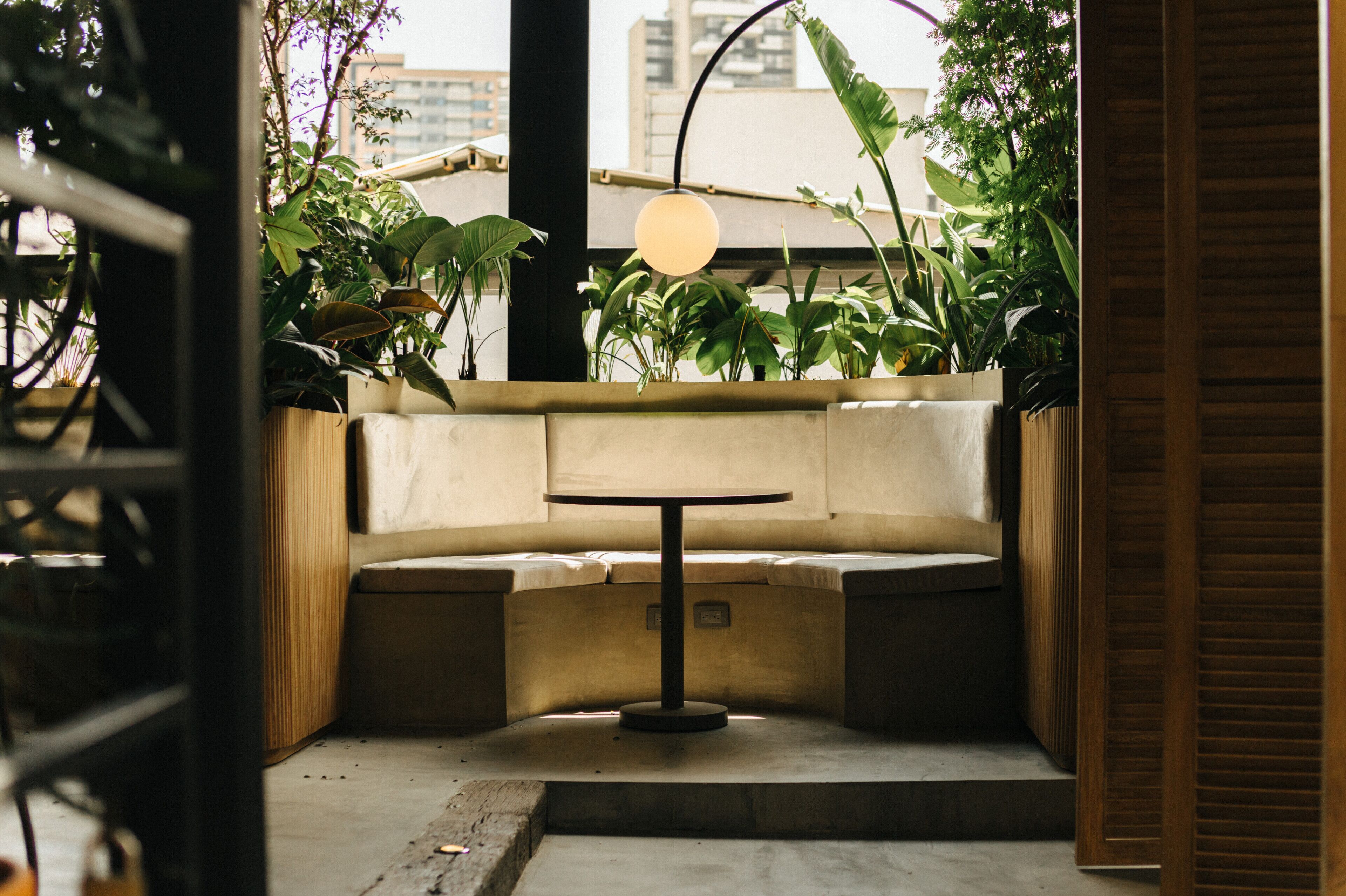 Uma área de estar com assentos em canto, com uma mesa redonda, rodeada por plantas em vasos e um lustre.