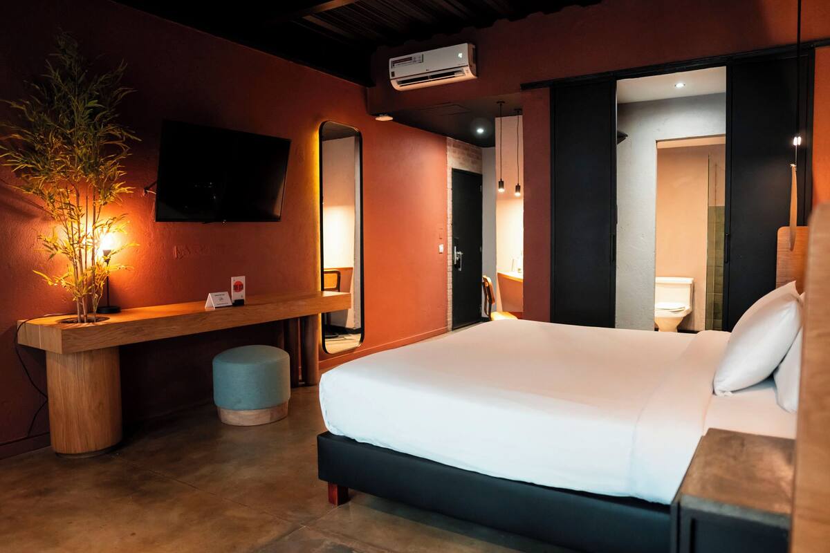 Habitación de hotel con cama, escritorio con silla, un espejo grande, equipo de aire acondicionado y una planta en maceta.