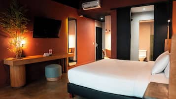 A hotel room with a bed, a desk with a chair, a large mirror, an air conditioning unit, and a potted plant.
