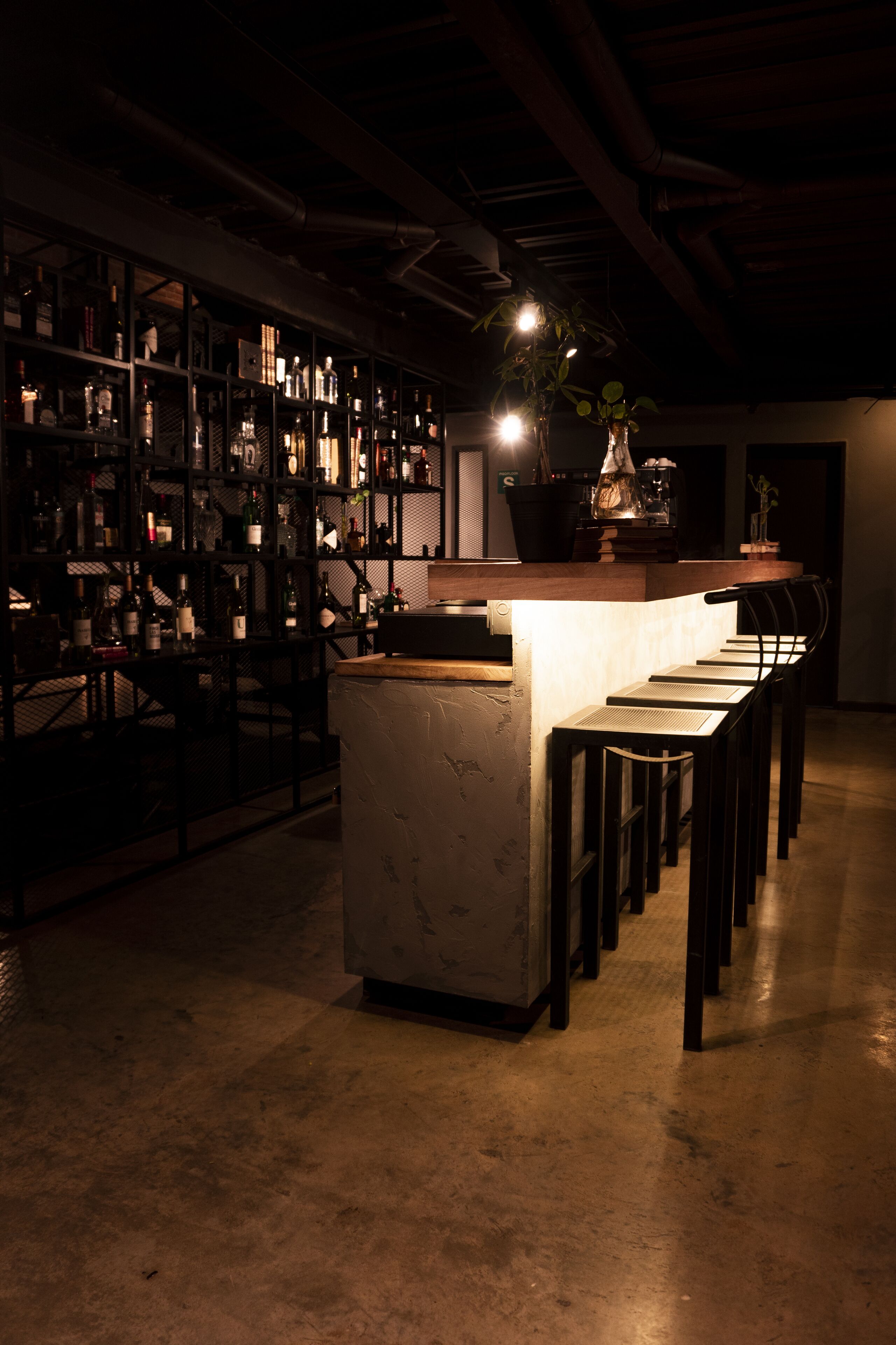 A bar area with a long counter and multiple stools. Shelves with bottles are visible in the background.