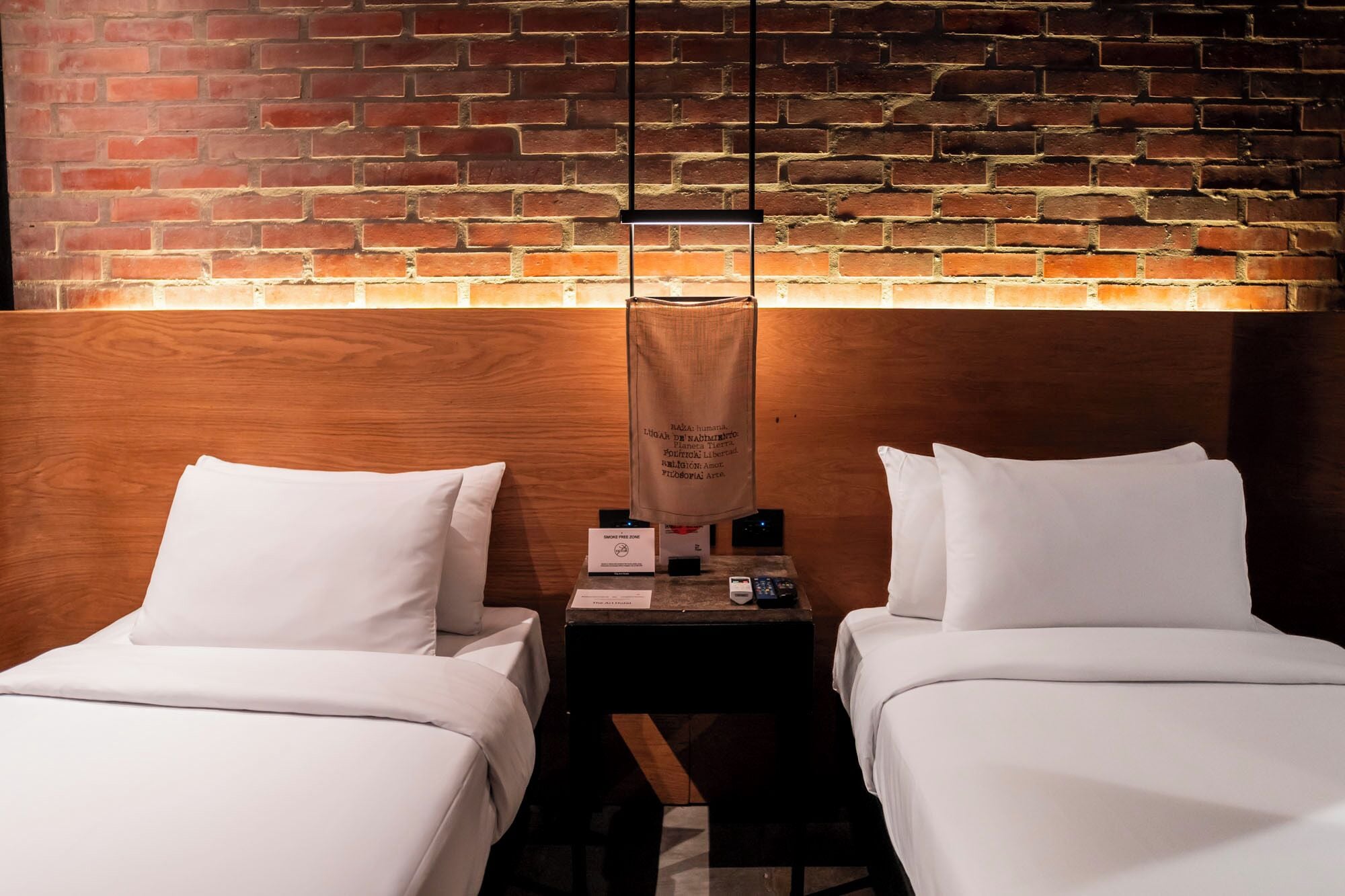 A hotel room with two beds, a brick wall, a wooden headboard, and a nightstand with a lamp and a sign.