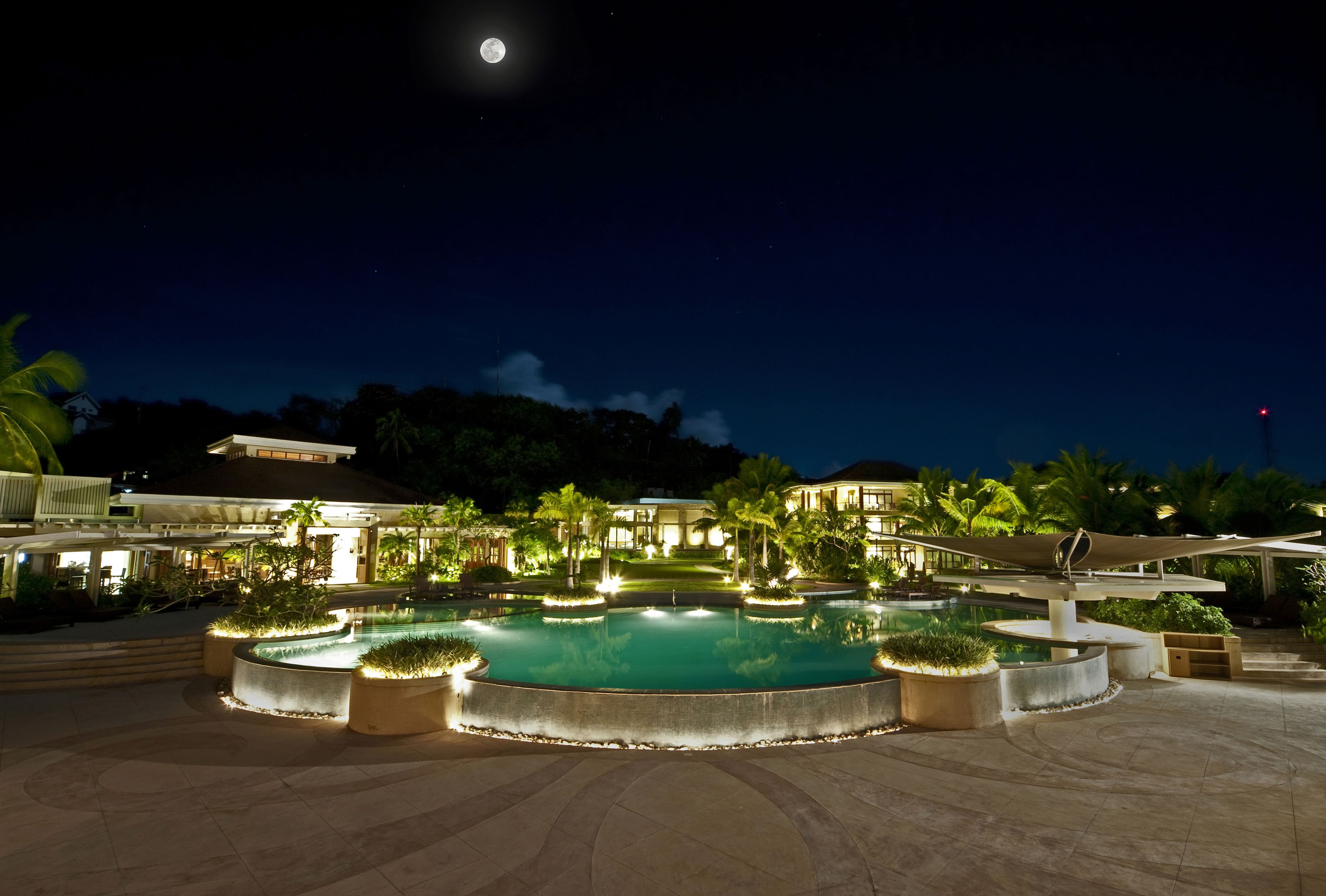 Zona de piscina nocturna con un elemento acuático circular, palmeras y un edificio al fondo.