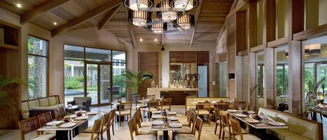 A spacious dining area with multiple tables and chairs, large windows offering views of the outdoors, and a central decorative light fixture.