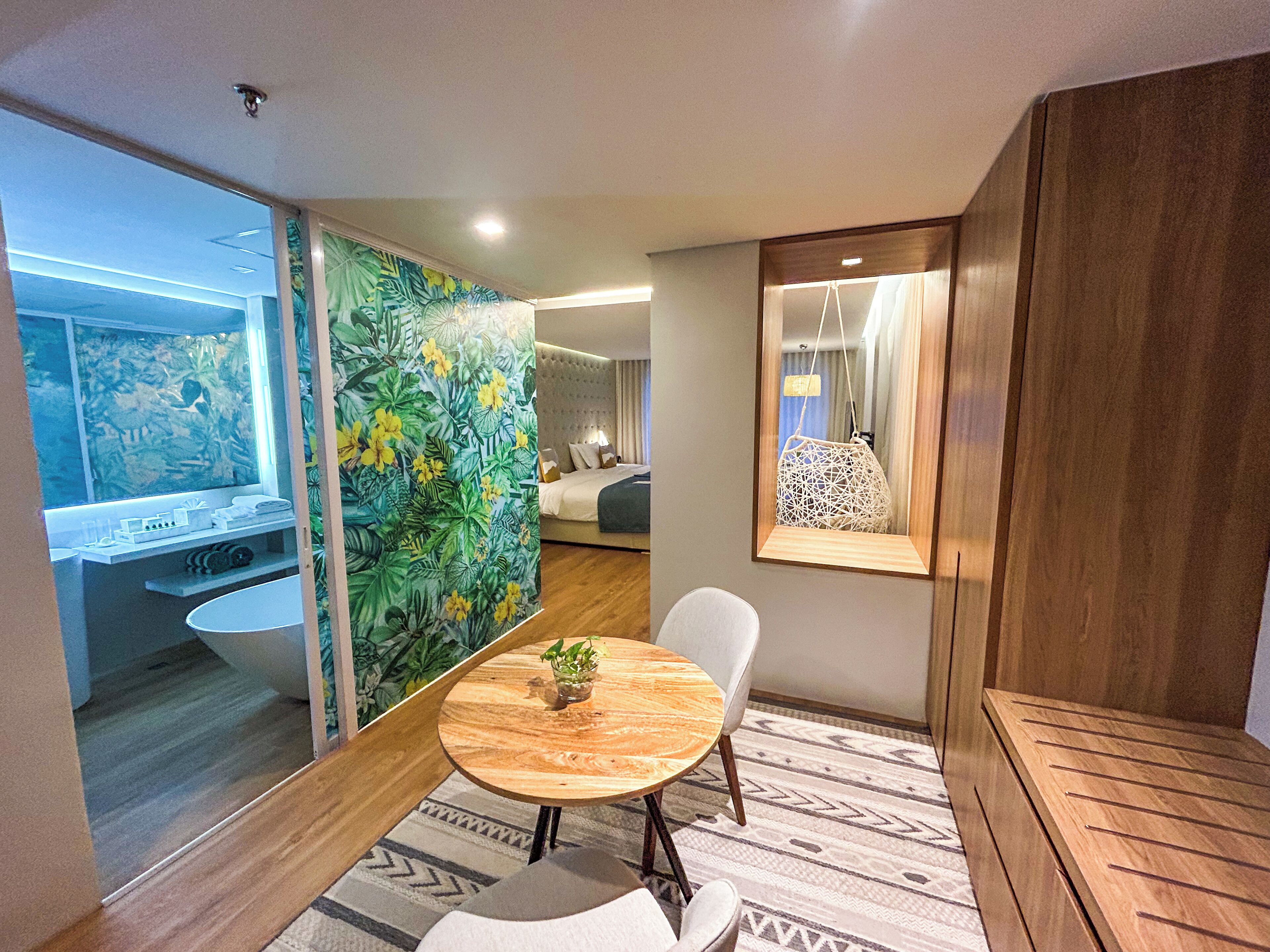 A modern hotel room with a bed, a round wooden table with chairs, a small potted plant, and a large mirror.