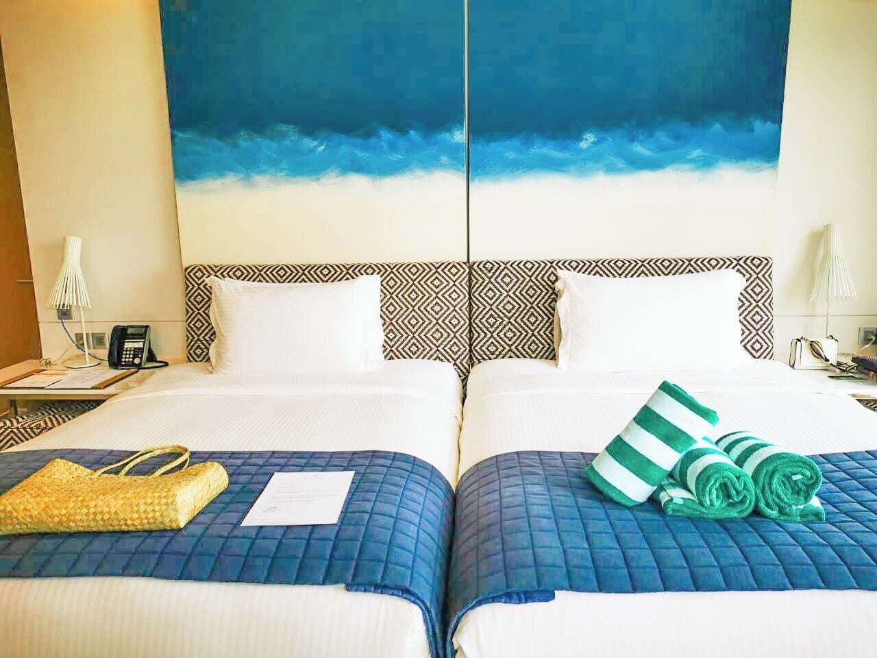 Ein Hotelzimmer mit zwei Betten, einem großen Gemälde eines blauen Himmels und Meeres sowie einem gemusterten Kopfteil.