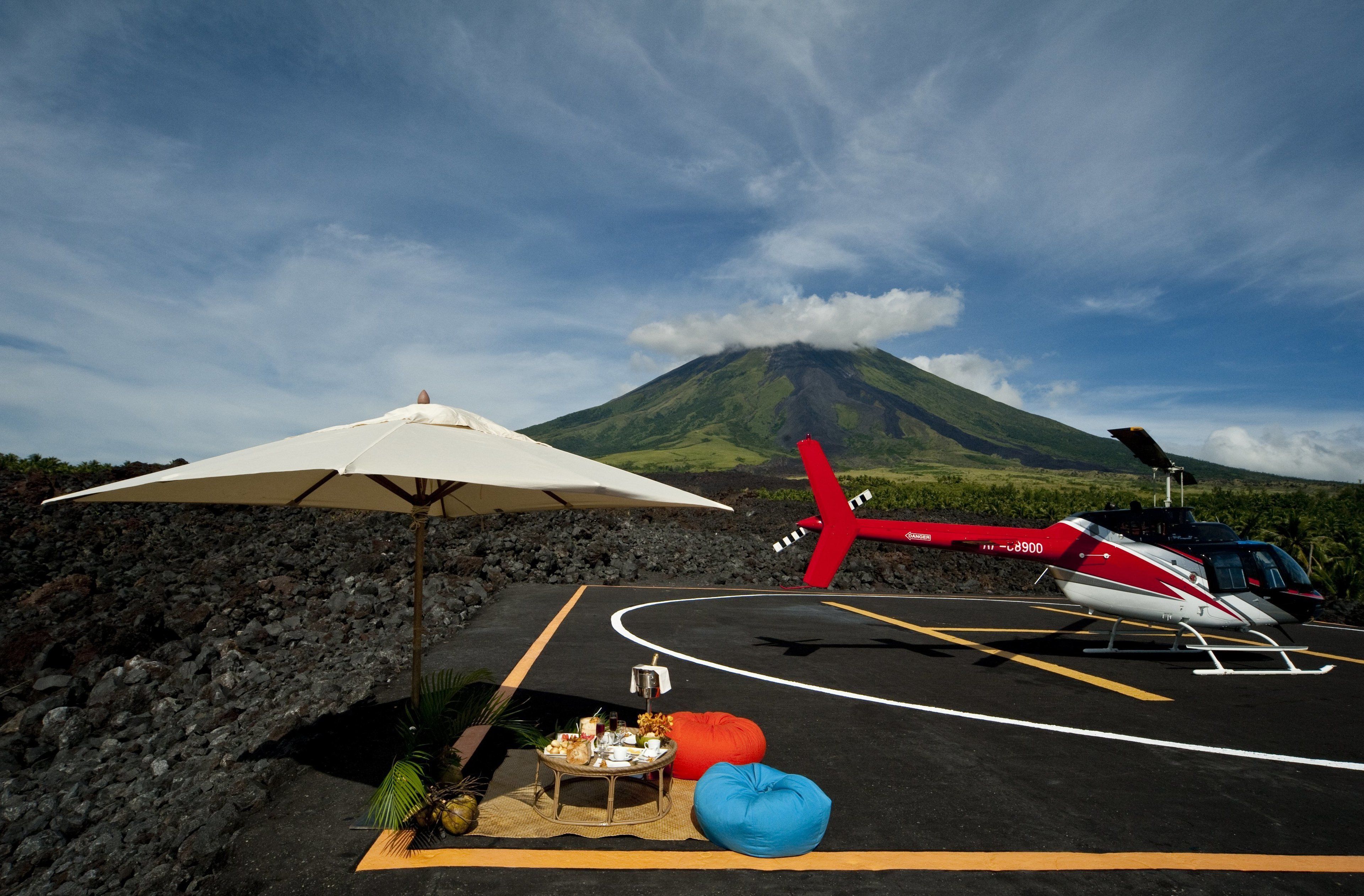 A helicopter on a helipad with a table and chairs, a red airplane model, and a mountain in the background.