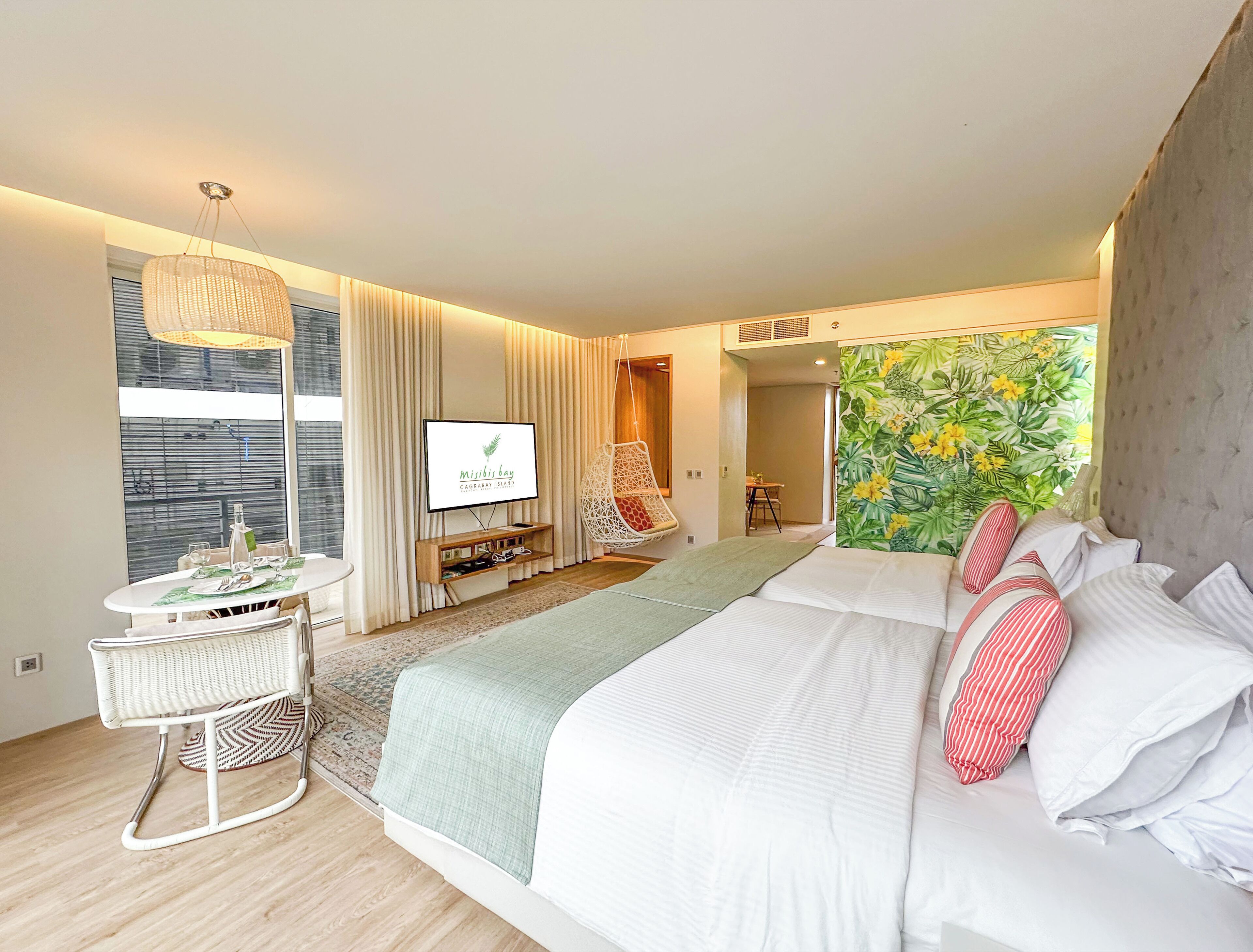 Habitación de hotel moderna con una cama grande, televisor, zona de comedor y decoración temática tropical.