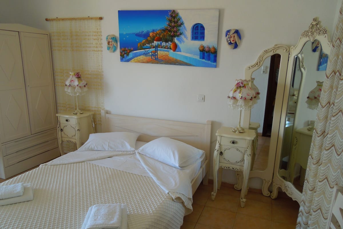 A neatly made bed with white linens, a painting of a coastal scene, and a mirror on a bedside table.