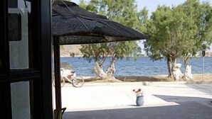 Vista desde una ventana a un patio con una mesa, sillas, una bicicleta y una masa de agua al fondo.