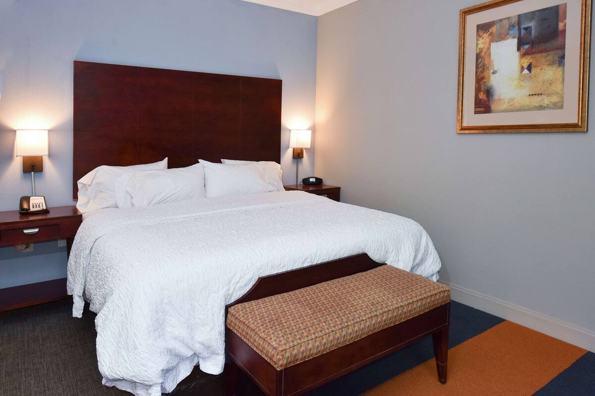 A neatly made bed with white linens, a wooden headboard, two bedside tables with lamps, a bench with a patterned cushion, and a framed abstract painting on the wall.