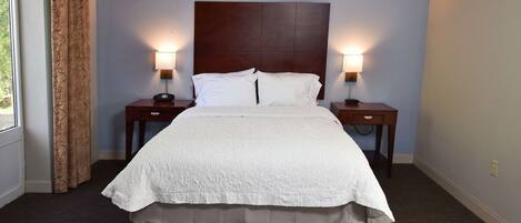 Una cama bien hecha con ropa blanca, flanqueada por dos mesitas de noche con lámparas, en una habitación con paredes azul claro y una ventana con cortinas.