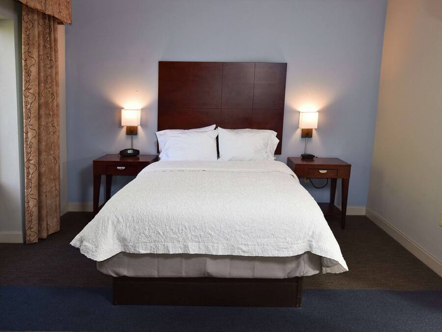 A neatly made bed with white linens, flanked by two bedside tables with lamps, in a room with light blue walls and a window with curtains.