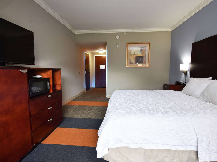A hotel room with a large bed, a flat-screen TV, a wooden dresser, and a carpeted hallway.
