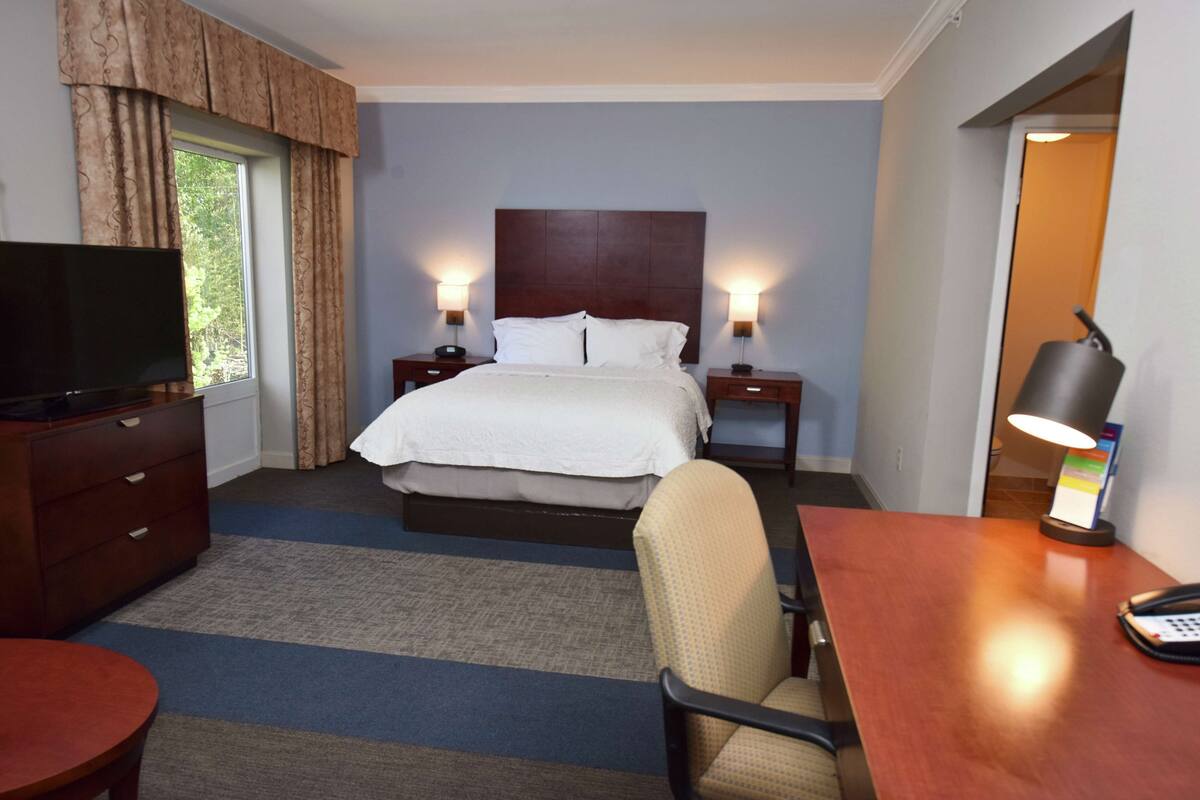 A hotel room with a bed, two bedside tables with lamps, a TV, a desk with a chair, and a window with curtains.