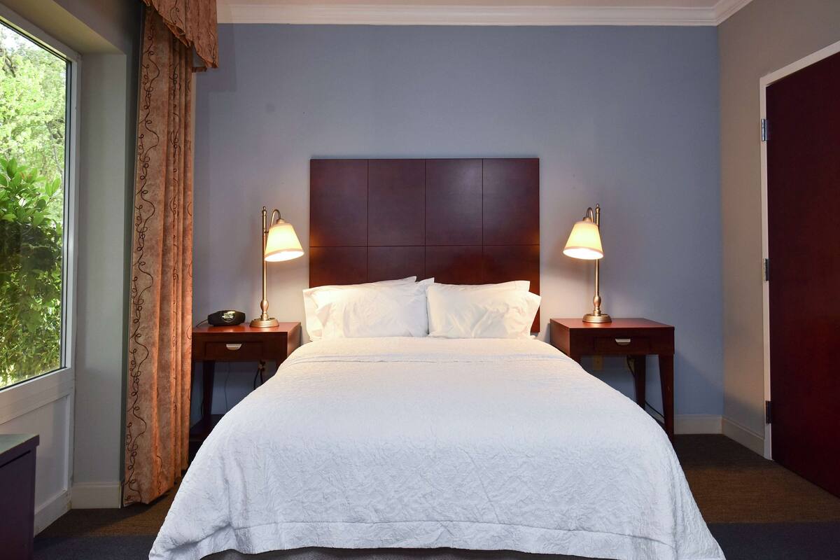 A neatly made bed with white linens, two bedside lamps, and a dark headboard in a room with a window and a door.