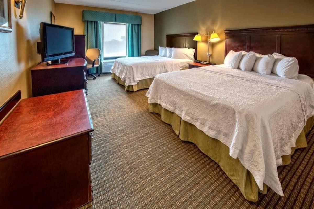 Une chambre d’hôtel avec deux lits, un bureau avec une télévision, une lampe et une fenêtre avec des rideaux.